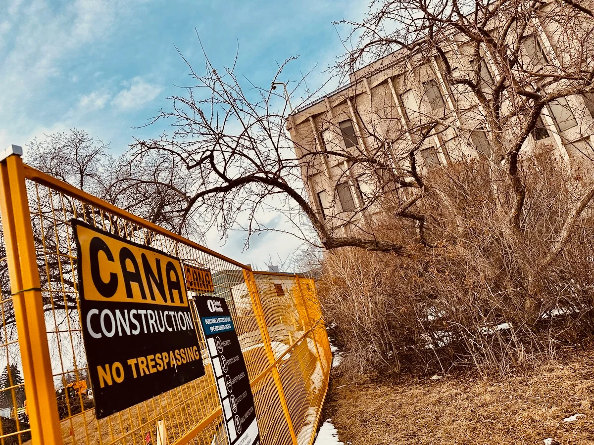 A construction site fence with a 'Cana Construction' warning sign indicating 'No Trespassing'. Behind the fence stands an abandoned or deteriorating concrete building surrounded by leafless trees and barren shrubs under a partly cloudy sky.