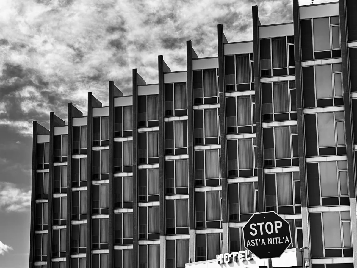 A black-and-white architectural shot looking up at a tall building with vertical columns and windowed balconies, set against a cloudy sky with a 'STOP' sign in the foreground.